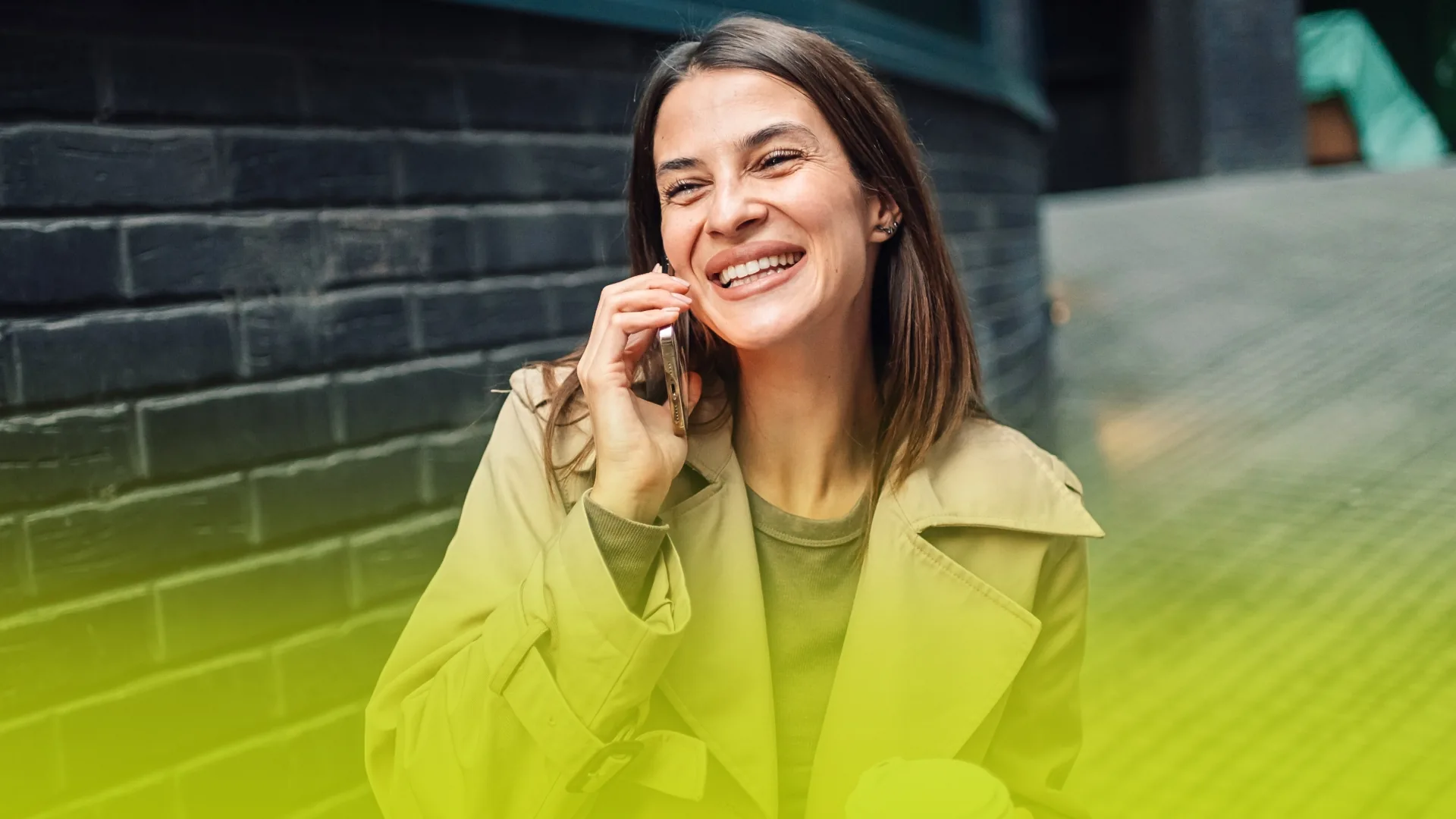 Woman on a phone call outdoors with a warm smile, neutral-toned coat, and urban brick wall background.