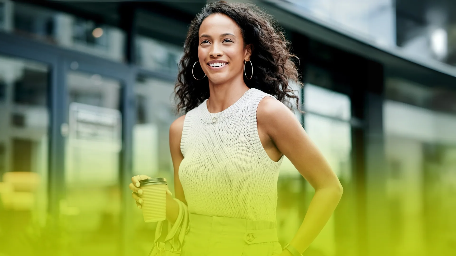 Woman with curly hair and hoop earrings carries a to-go coffee and handbag, sleeveless knit top, urban glass storefront behind her with yellow gradient.
